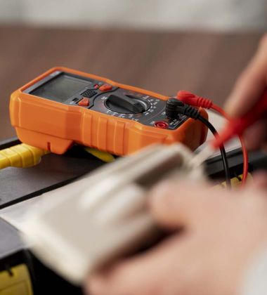 person checks a battery's voltage using an electronic multimeter in a well-lit workspace.