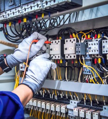 A person in blue overalls is repairing electrical equipment, focused on their task in a workshop setting.